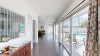Located at 1931 W 16th Ct in Riviera Beach, Florida, this single-floor space offers a well-organized layout that balances functional work areas with comfortable living. The open-concept kitchen and living room form the heart of the space, unified by rich, dark hardwood flooring that runs throughout. The kitchen is equipped with granite countertops, warm maple cabinetry with frosted glass upper panels, black appliances, and a peninsula that doubles as a breakfast bar. Pendant lighting adds a modern touch above the counters. The living area is bright and airy, with crisp white walls and large sliding glass doors that flood the room with natural light and provide direct views of an outdoor pool area. A dining area sits comfortably alongside the living space, making this an ideal setting for both relaxing and entertaining. A separate office provides a quiet, focused workspace, featuring a ceiling fan, a natural-light window, and ample room for a full workstation setup. Throughout the space, the clean white walls and open sightlines create a cohesive, welcoming atmosphere that reflects a practical yet modern Florida aesthetic.