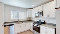 This well-organized multi-story home spans three floors and offers a thoughtful layout designed for comfortable everyday living. The ground floor provides direct access to an attached garage with a sectional door, alongside a welcoming entrance that leads to carpeted staircases connecting all levels. The main living floor features a bright, open-plan kitchen and living room. The kitchen is outfitted with white cabinetry, dark countertops, stainless steel appliances including a gas range and over-the-range microwave, and a single-basin sink beneath a window with natural light. The adjacent living area features light wood-look flooring, recessed ceiling lighting, and a clean, neutral palette. Upper-floor hallways lead to three carpeted bedrooms, each finished with white walls and blinds-covered windows. The generously sized primary bedroom includes an en-suite bathroom featuring a white vanity with an undermount sink, and a walk-in closet with dual hanging rods. A second full bathroom serves the remaining bedrooms. Throughout the home, consistent use of neutral tones, light wood flooring in common areas, and plush carpet in bedrooms creates a cohesive and move-in ready environment.