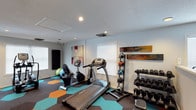 This fitness center offers a thoughtfully designed workout environment tailored to support a variety of exercise routines. The space is finished with a striking hexagonal-patterned floor in teal, orange, and charcoal tones, adding a vibrant, energetic atmosphere that complements the room's clean white walls and textured ceiling. Recessed lighting, combined with natural light streaming through multiple windows fitted with white blinds, ensures the space remains bright and welcoming throughout the day. Cardio equipment, including a treadmill and a stationary bike, is positioned toward the center of the room, offering ample space for comfortable use. A well-organized dumbbell rack lines one wall, stocked with a full range of free weights to accommodate strength training at multiple levels. A stability ball and additional accessories are available for flexibility and balance work. Decorative wall art in warm, abstract tones adds a creative touch to the space, elevating the overall aesthetic beyond a purely utilitarian environment. A wall-mounted pad provides space for stretching or martial arts practice. The layout is open and efficient, allowing multiple users to exercise simultaneously without feeling cramped, making this fitness center a functional and visually appealing space for daily workouts.