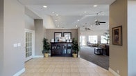 This single-floor space offers a thoughtfully designed interior that balances functionality with a welcoming ambiance. The reception and entry area greets visitors with light-toned ceramic tile flooring, vaulted ceilings with recessed lighting, and ceiling fans that contribute to an airy, comfortable environment. A dark wood display cabinet serves as a central focal point in the entryway, flanked by lush potted tropical plants that add a natural, refreshing touch to the interior.

The transition from the tiled entry into the adjoining lounge area is marked by a shift to patterned carpeting, delineating the more relaxed seating zone. The lounge is furnished with upholstered chairs and sofas arranged to encourage conversation and comfort. Large windows along the far wall allow generous amounts of natural light to fill the space, offering views to the outdoors and enhancing the open feel of the interior.

The neutral warm-gray wall tones throughout create a cohesive and calming aesthetic, while the vaulted ceiling adds a sense of vertical spaciousness. The overall design reflects a practical yet inviting layout well-suited for a community gathering or reception facility, prioritizing accessibility, comfort, and a clean, organized presentation.