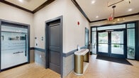 The Cloverleaf Gardens Clubhouse at 20 Foster Way in Woodbridge, NJ is a thoughtfully designed single-story community facility offering a comprehensive range of amenities. The entrance welcomes visitors through sleek double glass doors into a bright, herringbone-tiled foyer accented by wainscoting and a bold wall art installation, setting an upscale yet welcoming tone throughout.

The interior features two private offices with frosted glass partitions bearing the Cloverleaf branding, providing a professional environment for leasing consultations. Textured wall coverings, dark-trimmed doorways, and recessed lighting carry a cohesive modern aesthetic through the hallways and common areas.

The spacious fitness center is a standout amenity, outfitted with cardio machines including treadmills, an elliptical, and a stationary bike, as well as a full cable machine, free weights, benches, and a rowing machine. Vaulted ceilings, large windows overlooking greenery, and floor-to-ceiling mirrors enhance the airy, functional space.

Two accessible bathrooms feature dramatic dark hexagonal marble-look tile on both floors and walls, offering a sophisticated contrast to the rest of the facility. Outside, expansive landscaped grounds include a circular walking path, garden plantings, and open green space, completing this well-appointed community clubhouse.