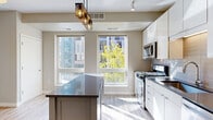 Located at 240 Chicago Ave in Minneapolis, MN, this one-bedroom, one-bathroom unit offers a thoughtfully designed single-floor layout that maximizes every square foot. The open-concept kitchen and living area serves as the heart of the home, featuring sleek white cabinetry that extends to the ceiling, stainless steel appliances including a gas range, microwave, and refrigerator, and a central island with a dark countertop that provides additional prep space and casual seating. A stylish pendant light fixture anchors the kitchen, while large windows flood the space with natural light and offer views of the surrounding Minneapolis cityscape and greenery. Light wood-style flooring runs throughout the main living areas, creating a warm and cohesive aesthetic. A hallway connects the living space to the private bedroom, which features soft carpeting and a ceiling fan, offering a quiet retreat. Two closets provide practical storage solutions. The full bathroom is finished with clean white tile surrounding the tub-shower combination, wood-look flooring, and a white vanity. Neutral warm-gray tones throughout the unit lend a modern, move-in-ready feel to this compact yet functional Minneapolis residence.