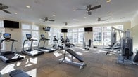 This single-floor fitness center is designed to provide a comprehensive workout environment in a bright, welcoming setting. Large windows line the walls, allowing abundant natural light to fill the space and offering views of the surrounding exterior. Multiple ceiling fans are mounted overhead alongside recessed lighting, ensuring consistent airflow and visibility throughout.

The cardio section features a row of treadmills and elliptical cross-trainers, each positioned to take advantage of the natural light and exterior views. Multiple flat-screen televisions are strategically mounted throughout the room, keeping members entertained during their workouts. The strength training area includes an adjustable weight bench, a shoulder/overhead press station, and a selection of free weights, providing options for a well-rounded resistance training routine.

The flooring consists of durable, patterned carpet designed to withstand high foot traffic while providing a degree of comfort underfoot. The neutral, light-toned walls contribute to the open and airy ambiance of the space. Overall, the fitness center is thoughtfully laid out to maximize usable floor space, offering a clean, functional, and inviting environment suited for a variety of exercise routines.