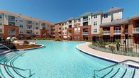 This outdoor amenity space is defined by its generously sized, freeform swimming pool with striking turquoise water, serving as the centerpiece of the community's exterior. The pool is bordered by a combination of smooth concrete and herringbone-patterned brick pavers, creating a polished, resort-like atmosphere. Rows of sleek lounge chairs are positioned along the poolside, offering ample space for sunbathing under open skies.

Adjacent to the pool, a covered pergola structure provides shaded outdoor living areas furnished with contemporary wicker sectional sofas and armchairs, accented by vibrant teal and orange cushions atop colorful area rugs. These seating areas are framed by lush landscaping, including ornamental shrubs and decorative trees, adding a natural softness to the hardscape surroundings.

Curved walkways connect the various zones of the outdoor space, leading residents seamlessly between the pool deck and lounge areas. The multi-story residential buildings surrounding the courtyard feature brick and siding exteriors with private balconies overlooking the amenity area. Iron fencing delineates the perimeter, maintaining a sense of privacy while preserving views of the landscaped grounds.