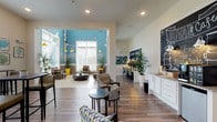 This single-floor community clubhouse at Mystic Point is thoughtfully designed to serve as a welcoming social and functional hub. The space opens into a generously sized main lounge area distinguished by soaring double-height ceilings, bold turquoise shiplap accent walls, and warm wood-look tile flooring that flows throughout. String lights and yellow pendant fixtures add a relaxed, festive ambiance, while an assortment of patterned lounge chairs and geometric coffee tables invite comfortable gathering. A striking stone fireplace anchors one corner of the lounge, complemented by lush tropical potted plants that reinforce the coastal aesthetic. Adjacent to the lounge is the Duval Café — a dedicated café station featuring white cabinetry, granite-style countertops, a beverage bar, and a large chalkboard wall welcoming residents to Mystic Point. Seating in this area includes a mix of high-top bar tables and stylish accent chairs. A separate dining nook is bathed in natural light from multiple large windows adorned with patterned valances, and decorated with coastal botanical artwork. A dedicated business center, framed by a wrought-iron railing, offers multiple computer workstations beneath a "Positive Vibes" chalkboard mural, providing a focused workspace within the communal setting. Together, these zones create a cohesive, resort-inspired environment designed for both productivity and relaxation.