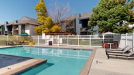 This outdoor amenity area centers around a generously sized, rectangular swimming pool with clear turquoise water, framed by a wide concrete pool deck that provides ample space for lounging and recreation. The pool features clearly marked depth indicators and is fully enclosed by a white fence, ensuring both safety and privacy for residents.

The deck is furnished with multiple reclining lounge chairs and a selection of colorful accent seating, offering a variety of relaxation options. A shaded seating area with a teal market umbrella adds a comfortable retreat from the sun. A wood-framed pergola structure is positioned adjacent to the pool area, creating an additional covered outdoor gathering space equipped with a grill — ideal for outdoor dining and social events.

A dedicated poolhouse building sits at one end of the pool, featuring large sliding glass doors and direct deck access. The structure provides functional support facilities for the pool area. In the background, distant mountain views are visible under clear blue skies, enhancing the open, airy atmosphere of the space.

Mature deciduous and evergreen trees surround the perimeter, providing natural shade and a lush, park-like setting. The multi-story residential buildings flanking the pool area feature blue-gray siding and private balconies that overlook the amenity space.