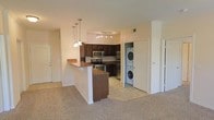 This single-floor residence showcases an open-concept layout that seamlessly connects the living area and kitchen. The kitchen is outfitted with rich, dark wood cabinetry, stainless steel appliances including a microwave, electric range, and dishwasher, and is illuminated by a track lighting system overhead. A peninsula-style breakfast bar with warm-toned countertops and decorative pendant lights serves as a natural divider between the kitchen and the carpeted living area, offering both additional prep space and casual seating. The living area features neutral wall tones and plush carpeting, contributing to a comfortable, cohesive atmosphere. A notable convenience feature is the in-unit stacked washer and dryer, neatly tucked into a dedicated closet space adjacent to the kitchen, eliminating the need for shared laundry facilities. Additional doors visible within the space suggest access to further rooms or storage. The overall design balances functionality with a clean, approachable aesthetic, making efficient use of the available layout throughout the single floor.
