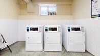 Located at 565 Avenue Rd in Toronto's Forest Hill neighbourhood, this shared laundry facility offers a well-organized, functional space designed to serve building residents efficiently. The room is finished with durable terrazzo flooring throughout, providing a clean and easy-to-maintain surface. Walls are painted in a neutral cream tone with white tile wainscoting in key areas, contributing to a bright and hygienic atmosphere. Overhead fluorescent strip lighting ensures the space is well-lit at all times.

The facility is equipped with multiple coin-operated commercial washers and dryers, including both front-load and top-load washing machines, as well as a row of side-by-side dryers positioned along one wall. A utility sink with built-in cabinetry provides added convenience for pre-treatment or hand-washing needs. A folding table and chairs are available, offering residents a comfortable area to sort and fold laundry. A cast iron radiator provides heat to the space, ensuring comfort year-round. Informational signage is posted on the walls, and a security camera is visible, reflecting attention to resident safety. The layout is open and efficiently organized to accommodate multiple users at once.