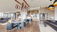 This elevated amenity space occupies a single floor high above the city, offering a refined blend of modern design and functional elegance. Throughout the space, rich herringbone-patterned hardwood flooring anchors each area, while floor-to-ceiling glass walls and oversized windows flood the interiors with natural light and frame panoramic urban skyline views.

The main lounge area is generously proportioned, accommodating multiple seating arrangements including plush sofas, accent chairs, and dining tables suited for both casual gatherings and organized events. Warm wood-toned built-in shelving units line the walls, displaying curated décor pieces and adding an inviting residential quality to the space. Striking sculptural pendant lighting—including distinctive ring chandeliers in both deep red and copper tones—serves as a bold focal point overhead, complemented by exposed wooden ceiling beams in select areas.

A full gourmet kitchen is outfitted with a large center island, gas cooktop, integrated appliances including a French door refrigerator and a dedicated wine cooler, and a dramatic veined stone backsplash that adds a luxurious touch. Open shelving provides ample display and storage.

An expansive wraparound outdoor terrace, accessible through sleek steel-framed glass doors, extends the living space outdoors with seating areas that capture sweeping city views, completing this sophisticated amenity floor.