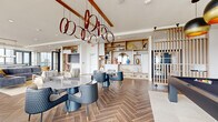 This elevated amenity space occupies a single floor high above the city, offering a refined blend of modern design and functional elegance. Throughout the space, rich herringbone-patterned hardwood flooring anchors each area, while floor-to-ceiling glass walls and oversized windows flood the interiors with natural light and frame panoramic urban skyline views.

The main lounge area is generously proportioned, accommodating multiple seating arrangements including plush sofas, accent chairs, and dining tables suited for both casual gatherings and organized events. Warm wood-toned built-in shelving units line the walls, displaying curated décor pieces and adding an inviting residential quality to the space. Striking sculptural pendant lighting—including distinctive ring chandeliers in both deep red and copper tones—serves as a bold focal point overhead, complemented by exposed wooden ceiling beams in select areas.

A full gourmet kitchen is outfitted with a large center island, gas cooktop, integrated appliances including a French door refrigerator and a dedicated wine cooler, and a dramatic veined stone backsplash that adds a luxurious touch. Open shelving provides ample display and storage.

An expansive wraparound outdoor terrace, accessible through sleek steel-framed glass doors, extends the living space outdoors with seating areas that capture sweeping city views, completing this sophisticated amenity floor.