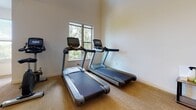 Located at 615 Rio Rd E in Charlottesville, Virginia, this well-equipped fitness center offers a comprehensive single-floor workout facility designed to support a wide range of exercise routines. The space features a clean, open layout with neutral-toned carpeted flooring, white walls accented by soft blue details, and bright overhead fluorescent lighting throughout.

The main exercise area is generously proportioned, accommodating multiple zones for cardio and strength training. Cardio equipment includes several treadmills and elliptical machines by Matrix, as well as stationary bikes, all positioned to take advantage of the large windows that frame views of surrounding greenery. Natural light flows in from multiple window groupings, creating an energizing and pleasant workout environment.

A dedicated strength training area features a multi-functional cable machine and a weight bench, while a dumbbell rack stocked with free weights provides additional resistance training options. High clerestory windows in one section of the space contribute to its notably tall ceiling height, enhancing the sense of openness. Structural columns are integrated throughout, delineating workout zones without disrupting the overall flow.

Access points include a windowed entry door and additional interior doors leading to utility and storage areas. Hand sanitizer stations are conveniently placed near the entrance, reflecting a health-conscious design. Overall, the facility provides a bright, well-organized, and functional environment for fitness at all levels.