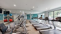 Located at 390 Stovall St SE in Atlanta, Georgia, this fitness center offers a well-appointed workout environment designed to support a range of exercise routines. The space is organized as a single open floor, providing ample room for both cardio and strength training activities.

The cardio zone is prominently featured along one side of the room, lined with multiple treadmills and elliptical machines, as well as a stationary bike. Full-length mirrors span the walls in this area, visually expanding the space and allowing users to monitor their form during workouts. Recessed lighting throughout the ceiling ensures a bright and energizing atmosphere.

On the opposite end, a dedicated strength and functional training area houses a multi-station cable machine, a medicine ball rack with an assortment of weighted balls, resistance tools, and exercise mats — providing versatile options for full-body conditioning.

A standout design feature is the bold teal accent wall emblazoned with the motivational phrase "JUST DON'T QUIT," serving as both a visual focal point and a source of inspiration. Large windows allow natural light to filter in, offering views of the surrounding greenery. The flooring transitions between carpet and a durable speckled surface depending on the zone, and a restroom is conveniently accessible within the space. The overall design is clean, modern, and purposefully functional.