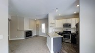 This single-story space showcases a thoughtfully designed open-concept layout that seamlessly connects the living area and kitchen. The living room is finished with plush carpeting and neutral gray walls, creating a warm and inviting atmosphere. A sleek fireplace with a clean white mantle and gray surround serves as a focal point, adding both style and function to the space. Natural light enters through a window near the front entry door, which features classic paneled detailing and white trim consistent throughout the interior.

The kitchen is well-appointed with a contrast of dark lower cabinetry and crisp white upper cabinets, complemented by a classic white subway tile backsplash. Stainless steel appliances, including a gas range and built-in microwave, add a modern touch. A peninsula with a light-toned countertop and an undermount sink provides additional workspace and casual seating potential. The kitchen area transitions to dark hardwood-style flooring, creating a clear and practical distinction from the carpeted living space. Recessed lighting throughout ensures the space is well-lit and functional.