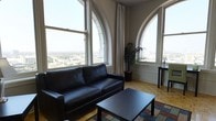 This remarkable space is defined by its striking architectural character, most notably the grand arched windows that dominate the living area and flood the interior with natural light. Set at a considerable elevation, the windows frame breathtaking panoramic views of the surrounding cityscape and beyond, creating a dramatic and ever-present connection to the urban environment outside. The living area is thoughtfully appointed, featuring a sleek black leather sofa and a coordinating coffee table resting atop a colorful area rug, all set against warm parquet hardwood flooring that adds a classic, timeless quality to the space. The arched window surrounds, finished in crisp white plasterwork, lend a sense of grandeur and historic architectural detail that elevates the overall ambiance. Sheer white curtains soften the abundant natural light while preserving the expansive views. A tall potted palm plant and a side table with a lamp add finishing touches that enhance the welcoming atmosphere. The combination of soaring architectural details, high-floor city vistas, and warm interior finishes creates a space that is both visually impressive and inviting — ideal for those who appreciate distinctive design and a vibrant urban backdrop.