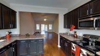 Located at 8254 County Rd 64 in Daphne, Alabama, this two-story home offers a well-designed open-concept layout that seamlessly connects the kitchen and living areas. The kitchen is a standout feature, appointed with rich dark cabinetry, granite countertops, and a central island that provides ample prep space and storage. Stainless steel appliances complement the modern aesthetic, while warm hardwood flooring flows continuously from the kitchen into the adjacent living and dining areas, creating a cohesive and inviting atmosphere. Crown molding adds a refined touch throughout the main living spaces. The living area is generously proportioned, anchored by a ceiling fan and bathed in natural light from multiple windows and a glass door that opens to the outdoors. The neutral wall tones enhance the bright, airy feel of the space. Spanning two floors, the home offers versatile living arrangements with room for comfortable everyday living and entertaining. The overall ambiance is warm and classic, combining traditional architectural details with updated finishes for a timeless residential appeal.