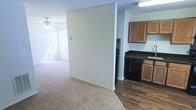 This single-floor space offers a practical and open layout that connects the living area to the kitchen. The living area is finished with neutral-toned carpet and features a ceiling fan with a light fixture, contributing to both comfort and air circulation. A window allows natural light to fill the space, creating a bright and airy atmosphere. The walls are painted in a clean, off-white tone throughout, lending a neutral backdrop that suits a variety of interior styles.

A partial wall divides the living area from the kitchen, providing a subtle separation while maintaining an open feel. The kitchen is distinguished by its dark hardwood-style flooring, which contrasts with the carpeted living space. Medium-toned wood cabinetry lines the upper and lower sections of the kitchen, accompanied by dark countertops. Black appliances, including a range and additional kitchen equipment, are neatly integrated into the layout. The textured ceiling is consistent throughout the space, adding a subtle visual detail overhead. Overall, the space presents a functional and straightforward design suited for everyday living.