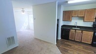This single-floor space offers a practical and open layout that connects the living area to the kitchen. The living area is finished with neutral-toned carpet and features a ceiling fan with a light fixture, contributing to both comfort and air circulation. A window allows natural light to fill the space, creating a bright and airy atmosphere. The walls are painted in a clean, off-white tone throughout, lending a neutral backdrop that suits a variety of interior styles.

A partial wall divides the living area from the kitchen, providing a subtle separation while maintaining an open feel. The kitchen is distinguished by its dark hardwood-style flooring, which contrasts with the carpeted living space. Medium-toned wood cabinetry lines the upper and lower sections of the kitchen, accompanied by dark countertops. Black appliances, including a range and additional kitchen equipment, are neatly integrated into the layout. The textured ceiling is consistent throughout the space, adding a subtle visual detail overhead. Overall, the space presents a functional and straightforward design suited for everyday living.