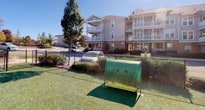 This single-floor residence is part of a well-established multi-story building community, offering both thoughtfully designed interior spaces and access to shared outdoor amenities. The exterior grounds feature a dedicated pet area enclosed by a chain-link fence, complete with artificial turf and a tunnel play structure, set against a backdrop of mature trees and a clear open sky.

Inside, the home is finished with dark hardwood-style flooring that extends through the hallways, connecting each room with a cohesive, polished aesthetic. The interior doors are classic two-panel white designs with brushed nickel hardware, complementing the neutral warm-beige wall tones throughout.

The bathroom is a standout feature, showcasing a granite countertop vanity with an undermount sink and brushed nickel fixtures, illuminated by a stylish multi-pendant vanity light bar. A large mirror enhances the sense of space. The full bathroom also includes a tub-shower combination finished with tasteful tile surround and a patterned shower curtain, along with a separate water closet for added privacy. Light tile flooring completes the bathroom's clean, contemporary look.

Overall, the space presents a clean, modern ambiance with quality finishes and community amenities that enhance everyday living.