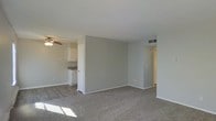 This single-floor space features an open-concept layout that connects the living area to the kitchen, creating a functional and cohesive flow throughout. The living area is finished with wall-to-wall carpet in a neutral gray-beige tone, contributing to a clean and understated aesthetic. Gray-painted walls and white trim run consistently throughout the space, lending a modern yet simple character to the interior.

A ceiling fan is positioned centrally in the living area, providing both lighting and air circulation. Natural light enters through windows, casting sunlight across the carpeted floor and brightening the space.

The kitchen is partially visible from the living area and features white cabinetry, dark countertops, and hard surface flooring that contrasts with the carpet of the adjacent room. A hallway extends toward the rear of the unit, suggesting additional rooms or storage spaces beyond what is immediately visible. Overhead HVAC vents are integrated into the walls, ensuring climate control throughout. The overall ambiance is clean, neutral, and move-in ready, offering a blank canvas for personalization.