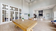 This impressive 830 square foot communal space in Orlando, FL, showcases a blend of modern design and functional elegance. The room's most striking feature is its soaring ceiling height, creating an airy and expansive atmosphere. Large windows and French doors line the walls, flooding the space with natural light and offering views of the surrounding area.

The open-concept layout is ideal for social gatherings and multifunctional use. The main area features a mix of seating options, including comfortable blue armchairs and wooden tables, perfect for casual meetings or relaxation. A prominent wooden table in the center serves as a focal point, suitable for group activities or dining.

The space includes a well-appointed kitchen area with dark wood cabinetry, granite countertops, and stainless steel appliances. A breakfast bar with modern stools provides additional seating and a casual dining option. State-of-the-art amenities such as wall-mounted televisions and a coffee station enhance the space's functionality.

The flooring throughout is a warm, light-toned wood, which complements the neutral wall colors and adds to the overall welcoming ambiance. Thoughtful lighting fixtures, including pendant lights and wall sconces, provide both task and ambient lighting options.

This versatile space seamlessly combines comfort, style, and practicality, making it an ideal setting for community gatherings, social events, or everyday use by residents.