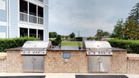 Located at 2222 Crow Ln in Myrtle Beach, South Carolina, this outdoor amenity space offers a well-appointed communal grilling station designed for convenience and entertaining. The centerpiece of the area is a pair of large stainless steel built-in grills, each equipped with multiple burners and lower cabinet storage, mounted on a sturdy brick base with a dark stone countertop that provides ample workspace for meal preparation. The clean, functional design reflects a commitment to quality outdoor living. Surrounding the grilling station is an open patio area with views of a manicured green lawn and mature trees, providing a pleasant and relaxed outdoor atmosphere. The adjacent multi-story residential building, finished in a coastal light-blue palette with white-railed balconies, frames the space and adds a charming, seaside character. The grilling station is well-positioned to serve residents and guests, making it an ideal spot for casual cookouts and social gatherings. The open sky and greenery beyond create a comfortable, airy setting that is characteristic of the Myrtle Beach coastal lifestyle.