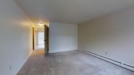This multi-story property spans three floors and presents a practical, no-frills layout suited for everyday living. The interior features wall-to-wall carpeting throughout the main living areas, providing a soft underfoot feel and a neutral aesthetic that serves as a blank canvas for personalization. Walls are finished in a clean, light beige tone that helps reflect ambient light and contributes to an open, airy atmosphere. Baseboard heating units are present along the walls, ensuring efficient and consistent warmth throughout the cooler months. The living area connects to a hallway that leads deeper into the home, hinting at a well-organized floor plan. Natural light filters in through vertical-blind-covered windows, brightening the space during daylight hours. Ceiling-mounted light fixtures provide additional illumination. Interior doors feature warm wood-toned finishes that add a touch of contrast to the lighter walls and flooring. The overall ambiance is clean and understated, with a functional design that prioritizes comfort and usability. The three-floor layout offers ample space distribution, making this property a versatile option for a variety of needs.