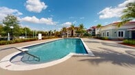 Located at 20950 NW 14th Pl in Miami Gardens, Florida, this single-level community amenity space offers a well-appointed outdoor environment designed for relaxation and recreation. At its centerpiece is a generously sized resort-style swimming pool with a smooth, light-colored deck that provides ample room for lounging, complemented by a separate spa for additional leisure. Tall, swaying palm trees and vibrant tropical plantings with red mulch landscaping beds frame the area, creating a lush South Florida atmosphere throughout. A shaded pergola structure near the pool offers a comfortable retreat from the sun, while classic wooden park benches are thoughtfully placed along the walkways for additional seating. A colorful children's playground with slides is situated at the far end of the grounds, making the space family-friendly. The main building features a distinctive Mediterranean-inspired architectural style, with a red barrel-tile roof and a soft sage-green exterior. Manicured shrubs and ornamental plants line the building's façade, enhancing its curb appeal. A gated entry provides security and privacy for the community. The entire outdoor space benefits from abundant natural sunlight and open sky views, embodying the vibrant and welcoming character of Miami Gardens living.