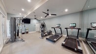 Located at 12 Wilkinson Way in Bluffton, South Carolina, this well-appointed fitness center offers a clean, comfortable environment designed to support a full range of workout routines. The space is finished with light, neutral-toned carpet flooring and soft blue-gray walls, creating a calm and focused atmosphere throughout.

The facility is equipped with multiple treadmills featuring integrated digital displays, an elliptical cross-trainer, and a multi-functional cable resistance machine that supports a wide variety of strength training exercises. A large wall-mounted mirror runs along one side of the room, visually expanding the space and allowing users to monitor their form during workouts.

Climate comfort is ensured by multiple sleek, black ceiling fans paired with recessed lighting, keeping the environment well-ventilated and evenly illuminated. A wall-mounted flat-screen television is positioned for easy viewing during cardio sessions. A convenience station stocked with cleaning supplies and fresh towels is available, reflecting a commitment to cleanliness and member experience.

A connecting hallway with light hardwood-style flooring links the fitness area to adjacent spaces, and a fire extinguisher cabinet is visibly mounted for safety compliance. The overall design is practical yet polished, making this fitness center a functional and inviting amenity.