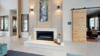 Located at 3 Woodcock Rd in Savannah, Georgia, this single-story community center for Ellis offers a welcoming and thoughtfully designed environment for residents and prospective tenants alike. The space makes an immediate impression at the entrance, where soaring ceilings, warm wood-framed floor-to-ceiling windows, and abundant natural light create an open, airy atmosphere. Lush potted grasses flank the glass entry doors, reinforcing a connection to the outdoors. The polished concrete flooring runs throughout, providing a sleek, modern foundation that ties the entire space together. A central lounge area anchors the interior, highlighted by a striking shiplap accent wall in a light wood finish that rises dramatically toward the ceiling. A contemporary linear fireplace with a clean stone surround serves as the focal point, flanked by wall-mounted accent lighting and nature-inspired artwork. A rustic sliding barn door adds a touch of warmth and textural contrast. Adjacent to the lounge, a live-edge wood communal table positioned beside large windows offers views of the outdoor pool area, making it an ideal spot for casual meetings or relaxed seating. An informational display area features a wall-mounted screen showcasing the Ellis community, along with brochures and signage. Access to fitness and restroom facilities is also conveniently located within the space.