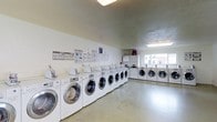 This shared laundry room is a practical, well-equipped facility designed to accommodate multiple users simultaneously. The space is lined with a generous row of front-loading washing machines along one wall, complemented by a corresponding set of dryers, providing ample capacity for residents. The machines are arranged in an organized, side-by-side configuration, maximizing the usable floor space while maintaining clear walkways throughout.

The room features smooth, light-colored walls that contribute to a clean, functional aesthetic. Overhead fluorescent lighting ensures the space is well-lit, while a window at the far end allows natural light to filter in, adding brightness to the environment. The polished concrete flooring is durable and easy to maintain, suited to the high-traffic nature of the space.

Informational charts and washing instruction guides are posted on the wall, offering helpful guidance for users. Additional storage cabinetry is visible toward the rear of the room. The overall layout is straightforward and utilitarian, prioritizing efficiency and ease of use, making it a convenient shared amenity.