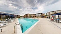 Located at 8501 Franklin Square Dr in Baltimore, Maryland, this outdoor amenity space is centered around a generously sized resort-style swimming pool with crystal-clear blue water. The pool is framed by a broad, open concrete deck that provides ample room for lounging, furnished with rows of chaise lounge chairs and umbrella-shaded seating areas in a coordinated blue and grey palette. A secure black iron perimeter fence encloses the pool area, ensuring both safety and defined boundaries for the space. An auxiliary structure on the deck serves as a poolside support facility, while additional covered seating areas nearby offer shade and social gathering spots. Surrounding the pool complex, the broader community grounds feature well-maintained lawns, mature trees, blooming ornamental plantings, and a paved pedestrian walkway that winds alongside the building exteriors. The residential buildings themselves are clad in a combination of stone veneer and contemporary siding, rising several stories with balconies overlooking the outdoor space. Large windows on the adjacent clubhouse-style building allow natural light to flow in and provide views of the pool area. The overall ambiance is that of a thoughtfully designed outdoor retreat, blending functional amenities with attractive landscaping to create an inviting communal environment.