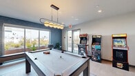 Located at 2050 Durant Ave in Berkeley, California, this contemporary game room is a well-appointed recreational space designed for entertainment and social gathering. The room is anchored by a full-size pool table positioned beneath a striking modern pendant light fixture featuring a row of globe-shaped bulbs on a sleek black frame, providing both functional illumination and a bold design statement.

A collection of classic upright arcade machines lines one wall, adding a vibrant, nostalgic touch to the space. Adjacent to the arcade area, a wall-mounted flat-screen television is paired with a built-in bench, creating a comfortable viewing area. Framed artwork decorates the surrounding walls, contributing to the room's polished aesthetic.

The space is finished with light-toned flooring and features a textured blue accent wall that adds depth and visual interest. Large sliding glass windows and a glass door open to a landscaped outdoor terrace, filling the interior with generous natural light and offering a seamless indoor-outdoor connection. Potted plants positioned throughout the room bring a refreshing touch of greenery. The overall ambiance is modern and inviting, balancing playful functionality with clean, contemporary design.