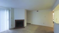 This single-floor space offers a straightforward and functional layout designed for comfortable living. The main living area is generously sized, featuring wall-to-wall carpet in a neutral beige tone that adds warmth underfoot. A standout feature of the room is the brick-surround fireplace, framed by a tiled hearth, which serves as a natural focal point and provides a classic, cozy aesthetic. The fireplace is flanked by a structural column that adds architectural character to the space. The walls are finished in a clean, neutral off-white, complemented by a textured popcorn ceiling that reflects the home's traditional construction style. Ventilation is provided through ceiling-mounted HVAC registers, ensuring efficient climate control throughout the area. The room benefits from an open, unobstructed layout that allows for flexible furniture arrangement to suit a variety of needs. Multiple electrical outlets are positioned along the baseboards for practical use. Overall, the space presents a classic residential interior with functional amenities and a neutral palette, providing an ideal canvas for personalization and interior design.