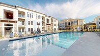 This outdoor pool area offers a well-appointed recreational retreat within a modern residential community. At its centerpiece is a generously sized swimming pool with crystal-clear blue water, framed by a smooth pool deck lined with lounge chairs — ideal for relaxing under open skies. The surrounding multi-story buildings feature classic architectural detailing with white and neutral-toned facades, large windows, and private balconies that overlook the pool area, creating a cohesive and attractive backdrop.

Adjacent to the pool is a covered pavilion structure finished in crisp white shiplap siding with coffered ceilings and sturdy decorative columns. This shaded space provides a comfortable gathering area directly connected to the pool deck, complete with potted greenery accents that add a touch of warmth. The pavilion also houses accessible restroom facilities, clearly marked for convenience, as well as a waste receptacle station for poolside cleanliness.

A gated black iron entry point connects the pavilion to surrounding walkways and green lawn areas, maintaining both security and a polished aesthetic. The overall design balances function and style, offering residents a clean, modern, and inviting outdoor amenity space suited for leisure and community enjoyment throughout the warmer months.