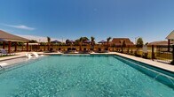 Located at 349 Ridge Rd in Lafayette, Louisiana, this outdoor community amenity space centers around a generously sized swimming pool designed for leisure and recreation. The pool area is thoughtfully laid out with wide concrete decking surrounding the water on all sides, providing ample room for lounging and movement. Multiple chaise lounge chairs are arranged along the pool's perimeter, accompanied by shade umbrellas that offer relief from the Louisiana sun.

A single-story clubhouse pavilion anchors one side of the pool area, featuring a hip-style shingled roof, white board-and-batten siding, and covered outdoor porches supported by dark wood columns. The covered patio areas provide additional shaded seating with wood tables and chairs, creating a comfortable gathering space. A patch of artificial turf adds a touch of greenery near the pavilion.

The pool area is enclosed by a black iron perimeter fence, ensuring both security and visibility to the surrounding residential neighborhood. Mature oak trees and newly planted palms and ornamental shrubs contribute to the natural ambiance. The broader community features single-story homes with classic architectural detailing, all visible under clear blue Louisiana skies.
