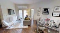 This single-floor residence showcases an open-concept layout that seamlessly connects the living and dining areas, creating a cohesive and functional living space. The interior is finished with warm hardwood-style flooring throughout, complemented by crisp white walls and tall baseboards that lend a clean, polished aesthetic.

The living area is anchored by a comfortable gray sofa accented with colorful throw pillows, paired with a sleek glass-and-chrome coffee table resting atop a plush white area rug. A wall-mounted flat-screen television is positioned opposite the seating arrangement, while framed artwork adorns the walls, adding character to the space. Glass-paned patio doors flood the room with natural light and provide access to the outdoors.

Adjacent to the living area, the dining space features a glass-topped table surrounded by contemporary upholstered chairs, ideal for entertaining. The open sightlines between both areas enhance the sense of spaciousness. A doorway visible in the background suggests additional rooms beyond, and the overall ambiance is bright, modern, and inviting, with a neutral palette that allows for versatile styling.