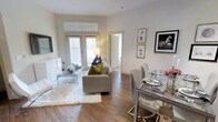 This single-floor residence showcases an open-concept layout that seamlessly connects the living and dining areas, creating a cohesive and functional living space. The interior is finished with warm hardwood-style flooring throughout, complemented by crisp white walls and tall baseboards that lend a clean, polished aesthetic.

The living area is anchored by a comfortable gray sofa accented with colorful throw pillows, paired with a sleek glass-and-chrome coffee table resting atop a plush white area rug. A wall-mounted flat-screen television is positioned opposite the seating arrangement, while framed artwork adorns the walls, adding character to the space. Glass-paned patio doors flood the room with natural light and provide access to the outdoors.

Adjacent to the living area, the dining space features a glass-topped table surrounded by contemporary upholstered chairs, ideal for entertaining. The open sightlines between both areas enhance the sense of spaciousness. A doorway visible in the background suggests additional rooms beyond, and the overall ambiance is bright, modern, and inviting, with a neutral palette that allows for versatile styling.
