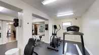Located at 4334 Appian Way W in Gahanna, Ohio, this single-floor fitness facility offers a dedicated space for exercise and wellness. The workout area is equipped with cardio machines, including a treadmill and elliptical trainer, providing residents or members with essential tools for a well-rounded fitness routine. A full-length wall mirror expands the sense of space while allowing users to monitor their form during workouts. The room features a textured ceiling with bright fluorescent lighting that ensures the space is well-lit throughout the day, complemented by a window that brings in natural light. The flooring is finished in a durable dark tone suited for high-traffic fitness use. An open pass-through connects to an adjacent mirrored area, which appears to offer additional equipment storage or a secondary workout zone, further enhancing the facility's versatility. The neutral white walls and clean layout contribute to a functional and focused atmosphere. Overall, this fitness space is thoughtfully arranged to maximize usability while maintaining a straightforward, practical environment for physical activity.