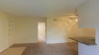 This single-floor residence offers a straightforward and functional layout designed for comfortable everyday living. The main living area is finished with wall-to-wall carpeting that lends a warm, understated feel to the space. Smooth, light-colored walls and a white ceiling contribute to a bright and airy atmosphere throughout. A ceiling fan is mounted overhead, providing both air circulation and a subtle lighting element. A central HVAC vent is visible on the wall, indicating a built-in climate control system. To one side, a countertop edge is partially visible, suggesting a transition into a kitchen or dining area finished with tile flooring — providing a practical contrast to the carpeted living space. A doorway leads to an adjoining room, which appears to benefit from its own natural light source, hinting at additional functional space such as a bedroom or utility area. Electrical outlets are distributed along the walls for everyday convenience. Overall, the space presents a clean, neutral palette with minimal ornamentation, making it well-suited for a variety of uses and easy to personalize.