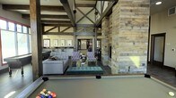 This single-floor space showcases a striking rustic-industrial aesthetic defined by soaring vaulted ceilings supported by exposed dark timber trusses and beams. A dominant reclaimed wood-clad column anchors the main gathering area, complemented by a built-in fireplace that adds warmth and character to the space. The open-concept layout is organized into several distinct zones. A recreation area in the foreground features a full-size billiards table, while an adjacent lounge area offers a gray sectional sofa and a tufted teal bench, creating a comfortable social gathering spot. Patterned accent chairs surround a central coffee table, adding a layer of visual interest. Further into the space, bar-height seating lines a counter area illuminated by overhead lighting. Framed artwork adorns the walls in the background, contributing to the polished yet approachable atmosphere. Dark tile flooring grounds the space and contrasts with the warm wood tones throughout. Natural light filters in from windows and exterior openings, enhancing the inviting ambiance. The overall design balances rustic charm with modern furnishings, making it a well-appointed and versatile communal space.