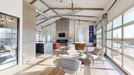 This thoughtfully designed single-floor space masterfully blends industrial and contemporary elements to create a dynamic, open-concept environment. Soaring vaulted ceilings with exposed wooden beams and visible industrial ductwork define the architectural character overhead, while warm hardwood flooring grounds the expansive interior below. Dramatic oversized glass garage doors line the walls, inviting an abundance of natural light and offering sweeping outdoor views that animate the space throughout the day.

The central living area serves as the heart of the space, anchored by a stunning floor-to-ceiling stone accent wall housing a sleek linear fireplace and a mounted flat-screen TV. A shiplap wood surround adds a refined rustic touch to this focal feature. Stylish pendant chandeliers hang at varying heights, complementing the recessed lighting to create a warm, layered ambiance.

The open layout connects multiple functional zones seamlessly, including a well-appointed wet bar with granite countertops and stainless steel appliances, a casual lounge area with modern seating, and a games zone complete with a shuffleboard table. French doors provide additional access points to the outdoors. Hallways connect supporting areas and ensure smooth circulation throughout. The overall atmosphere is bright, airy, and sophisticated — ideal for social gatherings and community interaction.