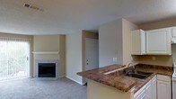 This single-story space is designed with an open-concept layout that seamlessly connects the kitchen and living areas. The kitchen features a raised bar counter finished with rich, granite-style countertops in warm earth tones, providing both a functional workspace and a natural dividing point between the two areas. Upper cabinetry in a light finish offers ample storage, while a stainless steel sink is conveniently positioned along the counter. The living area is carpeted in a neutral tone and centers around a tiled fireplace with a clean white mantel, serving as a natural focal point. The fireplace surround features classic brick-style tile detailing. Walls throughout are finished in a warm beige, complemented by a textured ceiling. A white paneled door provides entry access, flanked by open wall space that lends a clean, uncluttered feel. Ceiling vents are integrated discreetly, suggesting a central HVAC system. The overall ambiance is functional and understated, with a straightforward design that emphasizes practicality and comfort in a cohesive, open living environment.