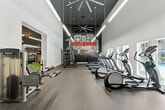 Located at 2235 Treehouse Ln in Corona, CA, this fitness center spans a generously sized single-floor layout designed to accommodate a wide range of workout disciplines. The main gym area features soaring ceilings with large ceiling fans and sleek linear pendant lighting, creating a bright and energizing atmosphere. A bold motivational mural anchors one end of the room, displaying fitness-themed typography in a striking black-and-red design. This area is outfitted with a row of treadmills and elliptical machines positioned along a wall of windows that offer views of an outdoor pool, allowing natural light to flood the space. A dedicated strength training zone includes a multi-station cable machine, free weights, barbells, and weight plates. An adjoining functional training area features artificial turf flooring and a TRX suspension training rig, complemented by a scenic mountain mural backdrop. A separate spin studio with warm hardwood flooring and high ceilings houses multiple Spinning-brand stationary bikes, providing an intimate space for cycling workouts. Throughout the facility, durable rubber flooring in the main zones ensures safety and comfort. The overall design balances industrial-modern aesthetics with practical, performance-focused functionality.