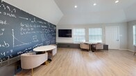 Located at 1800 Holleman Dr in College Station, TX, this thoughtfully designed communal space seamlessly blends study functionality with a comfortable lounge atmosphere. The single-floor layout is open and well-organized, making efficient use of its compact footprint.

The space is anchored by a striking full-wall chalkboard mural covered in scientific and mathematical equations, lending an academic character well-suited to its College Station setting. Vaulted ceilings in the lounge area add a sense of openness and volume, while recessed lighting and large windows fitted with sheer roller shades allow generous natural light to fill the room.

Light wood-tone flooring runs throughout, providing a warm, cohesive base that ties together both zones. The dining and collaborative work area features a high-top natural wood table paired with cognac leather bar-height chairs beneath a statement trio of matte black and brushed gold pendant lights. The lounge area offers multiple round pedestal tables with gold bases, each surrounded by plush blush velvet accent chairs — ideal for informal meetings or individual study. A wall-mounted flat-screen TV enhances the versatility of the space. Built-in low shelving along the walls provides additional surface area. The overall ambiance is modern, inviting, and academically inspired.
