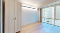 Located on the 7th floor at 145 Leavenworth Street in San Francisco's Tenderloin neighborhood, this efficiently designed unit offers a thoughtfully planned layout across a single floor. The interior embraces a clean, contemporary aesthetic with light hardwood flooring, crisp white walls, and a neutral palette throughout.

The kitchenette is outfitted with flat-panel cabinetry in a warm wood tone paired with dark upper cabinets, a marble-look countertop, a stainless steel sink with a matte black faucet, and built-in appliances including a microwave and dishwasher. Recessed under-cabinet lighting enhances both functionality and ambiance.

The bedroom is bright and open, benefiting from large floor-to-ceiling windows that invite natural light and offer urban views of the surrounding San Francisco cityscape. A wall-mounted flat-screen TV and built-in LED cove lighting add modern convenience, while a full-height wardrobe provides ample storage. The space is finished with the same light hardwood flooring found throughout.

The bathroom features an accessible, open-plan design with large-format white tile flooring, a wall-mounted sink, a wall-mounted toilet, a full-length mirror, and a roll-in shower equipped with matte black grab bars and a handheld showerhead. A mobile storage bench adds practical flexibility to the space.