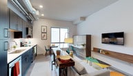 This thoughtfully designed studio showcases a single-floor, open-concept layout that maximizes every inch of its space. The industrial-chic aesthetic is immediately evident through the exposed ventilation ductwork running along the ceiling, which contrasts beautifully with the clean white walls and light hardwood flooring throughout.

The kitchen lines one wall with sleek navy-blue cabinetry, integrated appliances, and light countertops, offering a stylish and functional cooking area. Adjacent to the kitchen, a slim wooden console table serves double duty as both a dining surface and a transitional element between zones, complemented by teal Eames-style chairs that add a vibrant pop of color.

The living area is centered around a comfortable sofa, with a low-profile coffee table and a mounted flat-screen TV anchored to the wall. A dedicated workspace features a wall-mounted fold-out desk with walnut wood accents, paired with a modern chair — ideal for productivity without sacrificing floor space.

Natural light filters in through a large sliding window, brightening the entire space and creating an airy, open ambiance. Recessed ceiling lighting and a statement floor lamp provide warm, layered illumination throughout. Whimsical wall art adds personality, making this studio a functional yet inviting urban retreat.