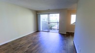 This single-floor space offers a clean, functional layout highlighted by rich, dark hardwood flooring that extends throughout the main living area, adding warmth and a sense of depth to the room. The walls are finished in a soft, neutral beige tone, creating a versatile backdrop suitable for a variety of interior styles. A textured ceiling adds subtle visual interest overhead.

A prominent sliding glass door serves as the focal point of the main living area, allowing generous natural light to fill the space and providing direct access to a private balcony enclosed by a dark metal railing. The outdoor area overlooks lush greenery, offering a pleasant connection to the outdoors. Sheer white curtains frame the sliding door, softening the transition between interior and exterior.

An additional window on the adjacent wall further enhances natural light, ensuring the space feels bright and open throughout the day. White baseboards provide clean definition along the perimeter of the room. The overall ambiance is straightforward and move-in ready, presenting a well-maintained, versatile living space with practical features and a comfortable, understated aesthetic.