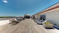 Located at 266 Morris Ave in Long Branch, New Jersey, this rooftop deck offers a generously proportioned outdoor retreat designed for both relaxation and social gatherings. The space is defined by wide-plank wood decking that extends across the entire rooftop, creating a warm and cohesive aesthetic throughout.

A standout feature is the partially covered lounge pavilion, anchored by a striking natural stone accent wall and finished with a beadboard ceiling equipped with ceiling fans for year-round comfort. This sheltered zone accommodates a large sectional sofa with deep blue cushions, complemented by a substantial outdoor dining table and chairs — ideal for al fresco entertaining.

Beyond the covered area, the open-air deck expands into a sun-drenched space furnished with lounge chairs and casual seating arrangements, perfect for soaking in the abundant natural light. Planters with greenery add organic texture throughout. Glass panel railings along the perimeter provide unobstructed panoramic views of the Long Branch skyline and surrounding neighborhoods, while maintaining a sleek, modern aesthetic. The thoughtful layout balances open exposure with sheltered retreats, making this rooftop an exceptionally versatile and inviting outdoor destination.