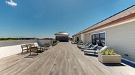 Located at 266 Morris Ave in Long Branch, New Jersey, this rooftop deck offers a generously proportioned outdoor retreat designed for both relaxation and social gatherings. The space is defined by wide-plank wood decking that extends across the entire rooftop, creating a warm and cohesive aesthetic throughout.

A standout feature is the partially covered lounge pavilion, anchored by a striking natural stone accent wall and finished with a beadboard ceiling equipped with ceiling fans for year-round comfort. This sheltered zone accommodates a large sectional sofa with deep blue cushions, complemented by a substantial outdoor dining table and chairs — ideal for al fresco entertaining.

Beyond the covered area, the open-air deck expands into a sun-drenched space furnished with lounge chairs and casual seating arrangements, perfect for soaking in the abundant natural light. Planters with greenery add organic texture throughout. Glass panel railings along the perimeter provide unobstructed panoramic views of the Long Branch skyline and surrounding neighborhoods, while maintaining a sleek, modern aesthetic. The thoughtful layout balances open exposure with sheltered retreats, making this rooftop an exceptionally versatile and inviting outdoor destination.