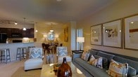 This single-floor residence offers an open-concept layout that integrates the living, dining, and kitchen areas into one cohesive and functional space. The living area is anchored by a plush gray sofa dressed with an assortment of patterned throw pillows, complemented by two white accent chairs featuring bold chevron cushions. A large decorative ceramic vase and a low coffee table serve as stylish focal points. Warm ambient lighting from table lamps and overhead pendant fixtures casts a golden tone throughout, enhancing the neutral yellow walls.

The dining area, situated just beyond the living space, features a compact table and chairs beneath a striking sunburst mirror and a decorative pendant light, creating an intimate setting for meals. Framed artwork adorns the walls, contributing to the polished, curated aesthetic of the space. A partial view into the kitchen reveals dark cabinetry and pendant lighting, suggesting a well-appointed culinary area. The seamless flow between zones, combined with consistent warm tones and carefully chosen furnishings, gives the overall space an elegant yet comfortable ambiance.